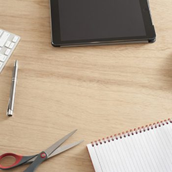 High Angle View of Desk with Scattered Office Supplies and Copy Space - Computer Keyboard and Mouse, Tablet, Scissors, Pen and Notebook on Wooden Desk with Central Blank Space